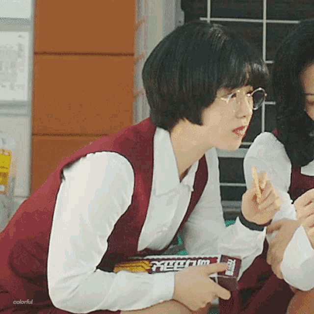 a girl wearing glasses and a red vest is holding a candy bar .