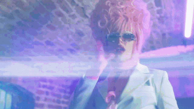 a woman with pink hair and sunglasses is standing in front of a brick wall .