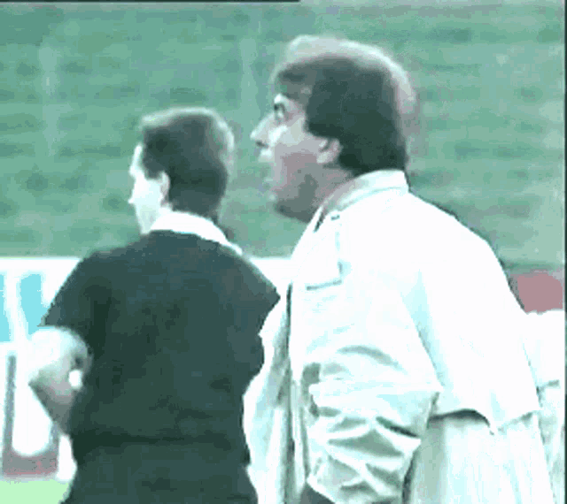a man in a white jacket stands next to a man in a black shirt on a soccer field