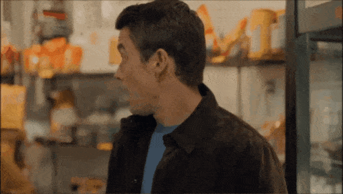 a man wearing a blue shirt and a brown jacket stands in front of a shelf full of food