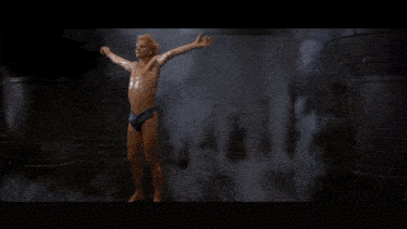 a shirtless man with red hair and blue eyes is smiling in a dark room