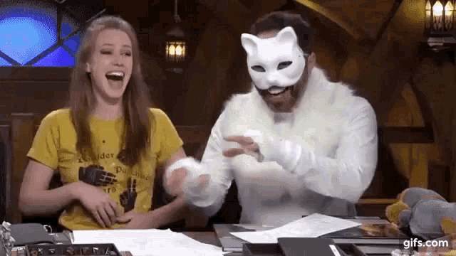 a man and a woman are sitting at a table laughing while the man is wearing a cat mask .