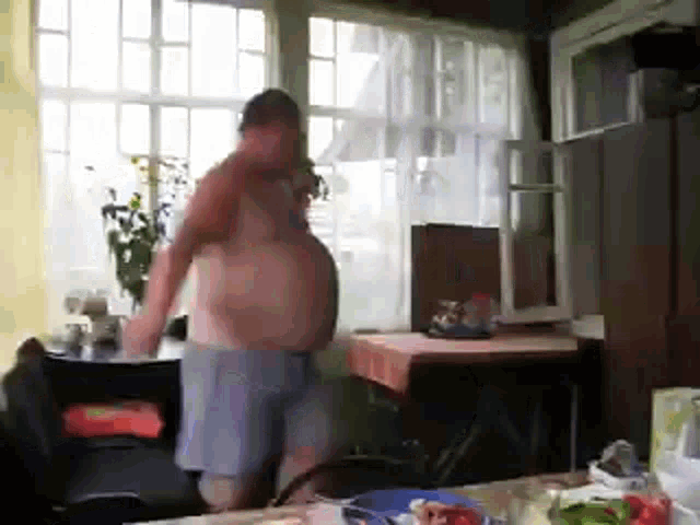 a shirtless man is standing in front of a window in a room with a table and chairs .