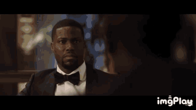a man in a tuxedo is looking at another man in a dark room with imgplay at the bottom of the screen