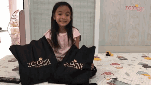 a little girl is sitting on a bed holding two bags that say zaracute