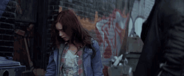 a woman in a blue jacket stands in front of a brick wall that has graffiti on it