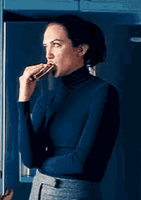 a woman in a turtleneck is eating a sandwich in front of a refrigerator .