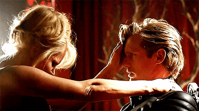 a woman is touching a man 's forehead in a room with a red curtain behind them