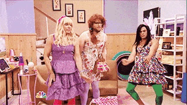 three people in dresses are dancing in a living room with a nbc logo on the wall