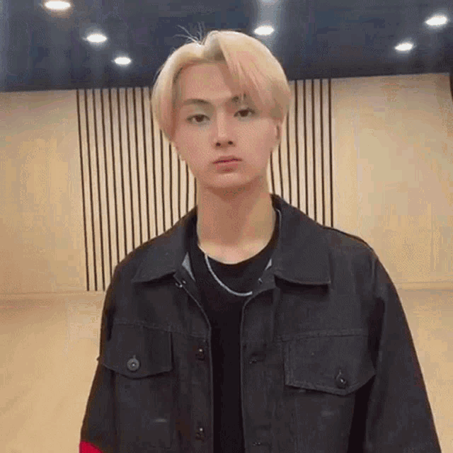 a young man with blonde hair is standing in a room wearing a black jacket and a black shirt .
