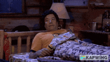 a man in a yellow shirt is laying in a bed with a kapwing logo on the bottom right