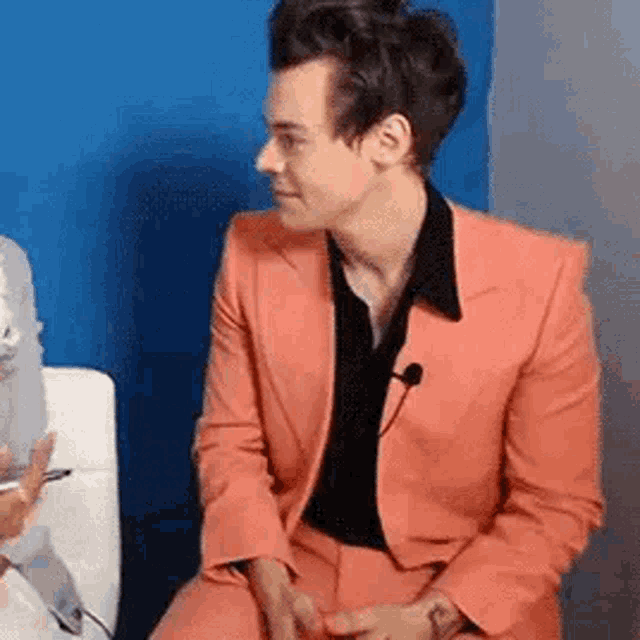 harry styles is wearing a pink suit and black shirt while sitting on a chair .