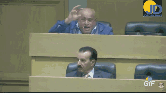 a man in a blue jacket is giving a speech in front of a screen that says jordan tv