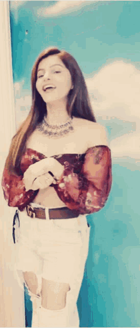 a woman wearing a red off the shoulder top and white jeans stands in front of a blue wall