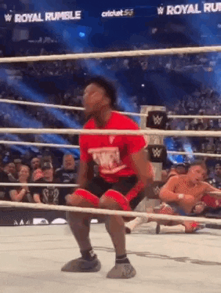 a man in a red shirt is kneeling in a wrestling ring while another man is laying on the ground .