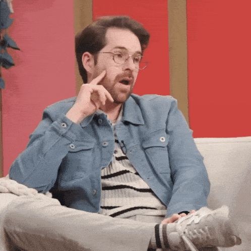a man wearing glasses and a denim jacket is sitting on a couch