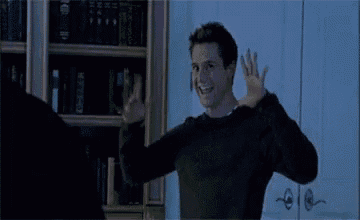 a man in a black sweater is standing in front of a bookshelf with his arms in the air .