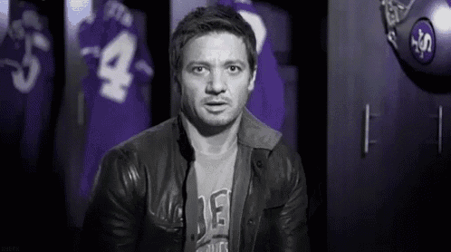 a man in a leather jacket and a t-shirt is standing in a locker room with purple jerseys .