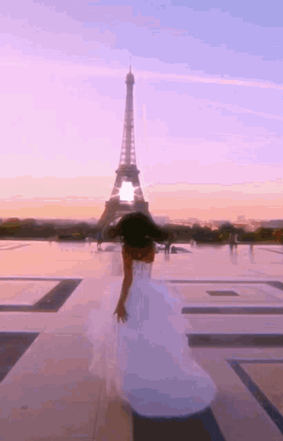 a woman in a white dress is standing in front of the eiffel tower at sunset
