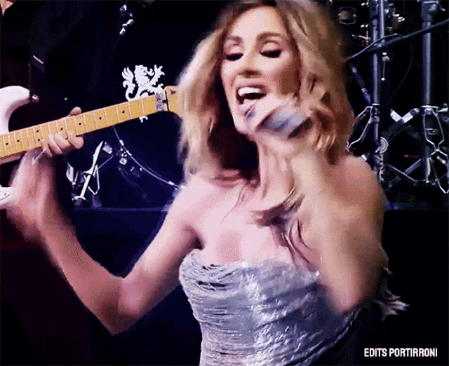a woman in a silver dress is singing into a microphone while holding a guitar behind her