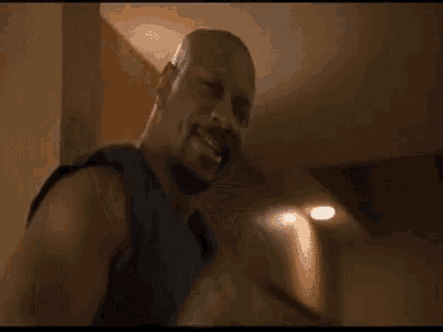 a man in a black tank top is standing in a room with a light on the wall .
