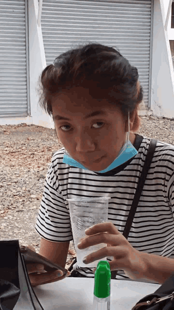 a woman wearing a striped shirt and a blue mask holds a cup of water