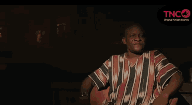 a man in a striped shirt is sitting in a dark room with a tnc logo behind him