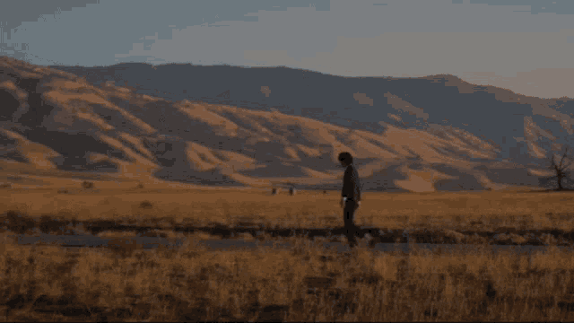 a man in a suit walks across a field with mountains in the background