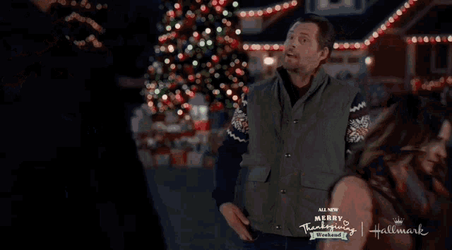 a man and a woman are standing in front of a christmas tree and a sign that says merry thanksgiving