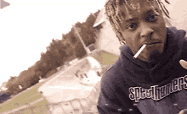 a man with dreadlocks is smoking a cigarette and wearing a specsavers hoodie .
