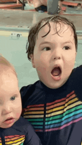 a boy and a baby are sitting next to each other in a swimming pool and the baby is making a surprised face .