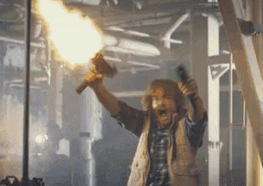 a man in a plaid vest is holding a gun with flames coming out of it 's barrel .