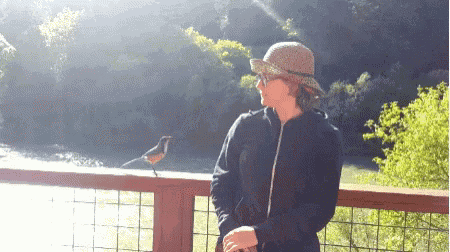 a woman in a hat stands next to a bird on a railing overlooking a river