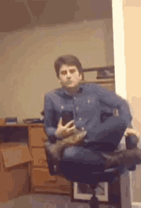 a man in a blue shirt is sitting in an office chair with his legs crossed