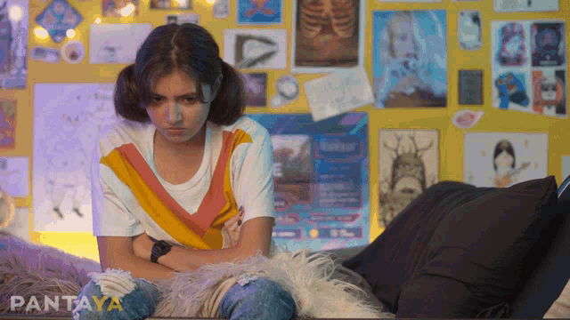 a girl is sitting on a couch with her arms crossed in front of a wall with posters on it that says pantaya