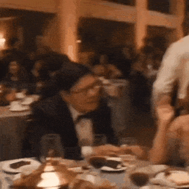 a man in a tuxedo is sitting at a table in a restaurant with glasses of wine .