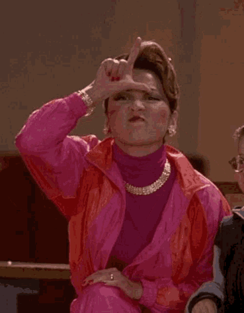 a woman in a pink jacket and orange pants is making a funny face with her hand .