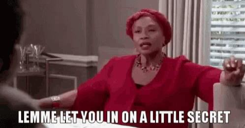 a woman in a red dress is sitting at a table talking to a man and telling him to let her in on a little secret