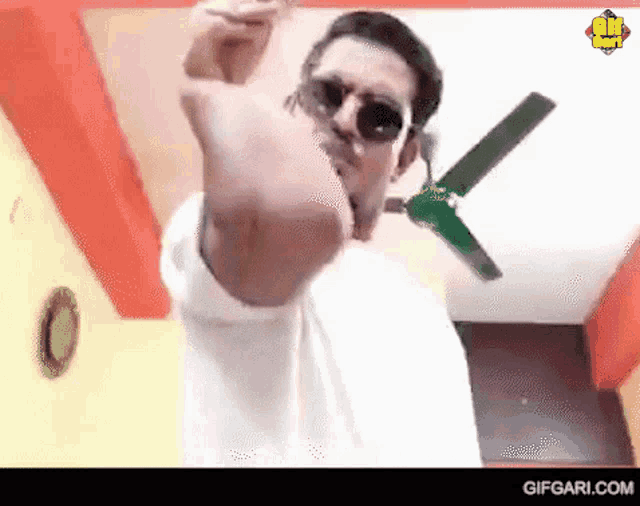a man wearing sunglasses is standing in front of a ceiling fan and giving the middle finger .