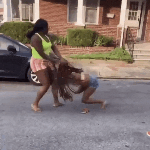 two women are fighting on the side of the road in front of a brick house .