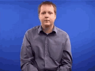 a man in a blue shirt is standing in front of a blue background and making a gesture with his hands .