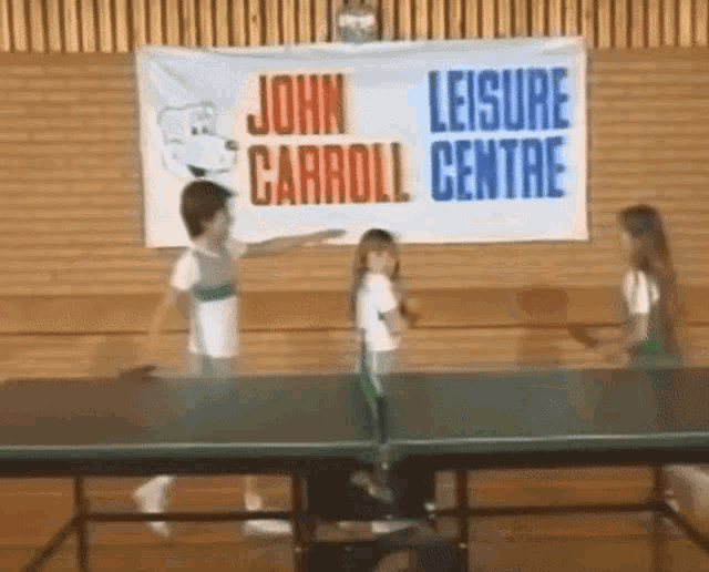 three young girls are playing ping pong in front of a john carroll centre banner