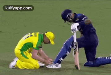 a cricket player is kneeling down next to another cricket player who is tying his shoe .