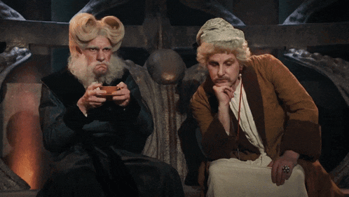 a man with a beard sits next to another man with a wig