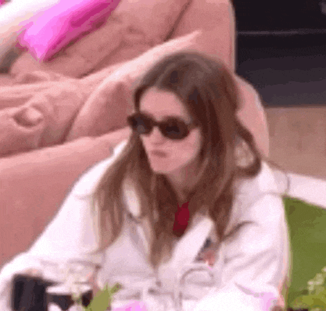 a woman wearing sunglasses and a white coat is sitting on a pink couch .
