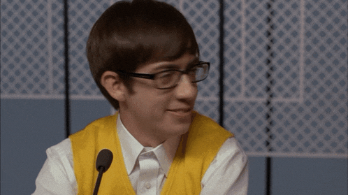 a young man wearing glasses and a yellow vest is smiling in front of a microphone
