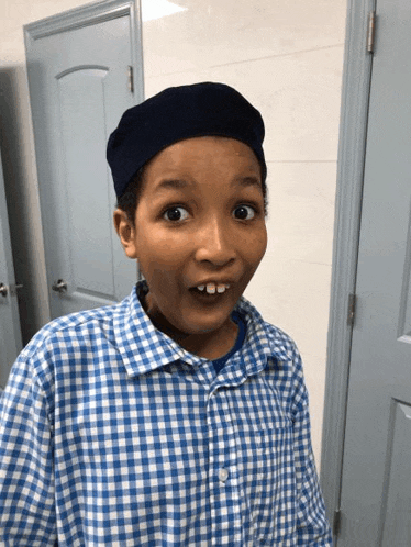 a young boy wearing a blue hat and a blue and white checkered shirt is making a funny face