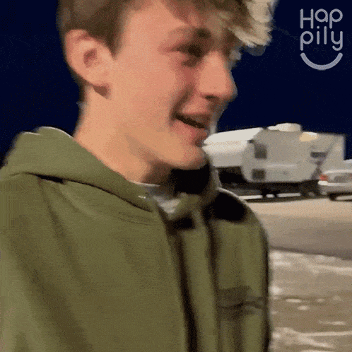 a young man wearing a green hoodie is smiling in front of a camper trailer .
