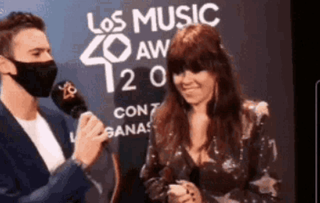 a man wearing a mask is interviewing a woman in front of a sign that says los music 40 aw