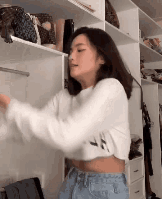 a woman in a white crop top is standing in front of a closet full of handbags .